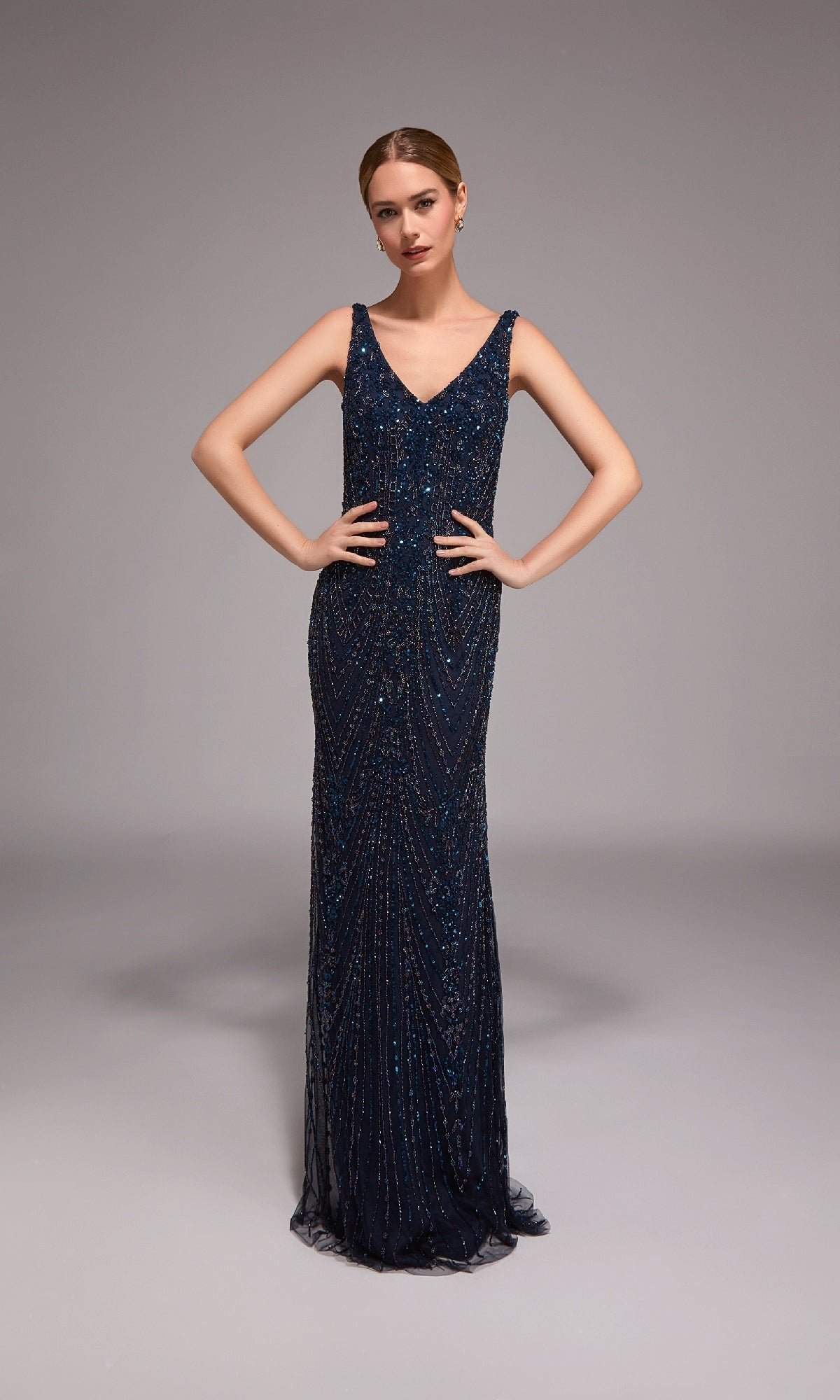 A woman poses in a studio wearing the Alyce 27786 long formal dress, a navy sleeveless beaded column gown with a V-neckline and floor-length design, her hands resting on her hips.